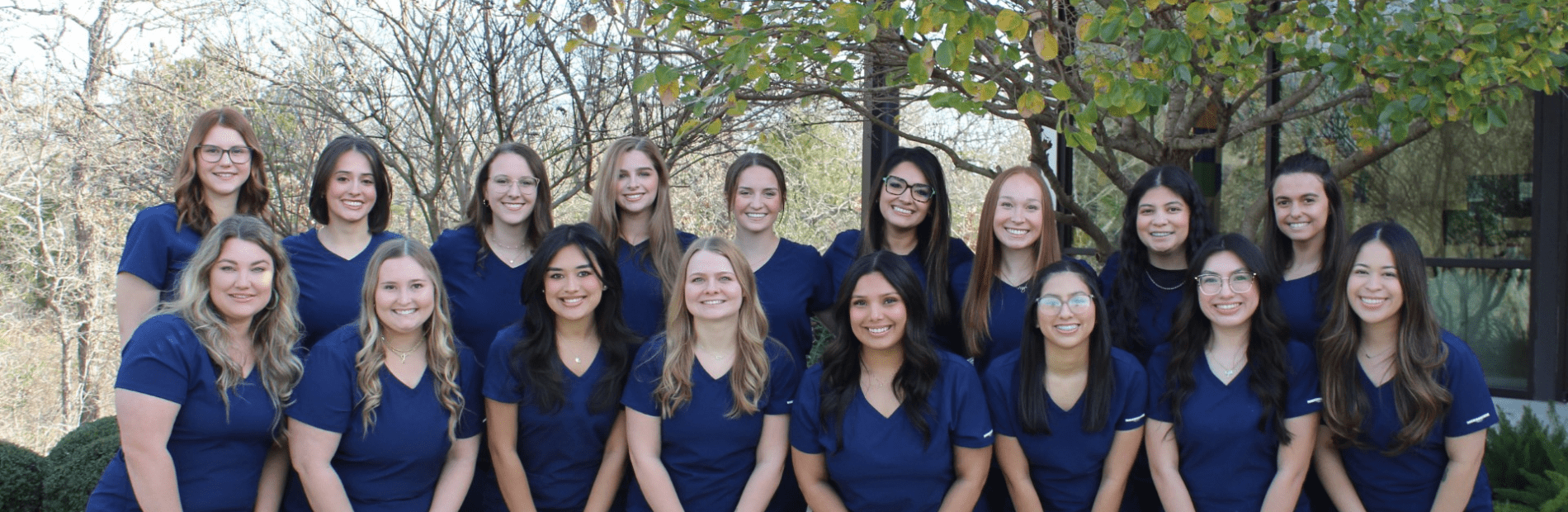 A group of seventeen women wearing navy blue scrubs poses outdoors in front of trees and a building.