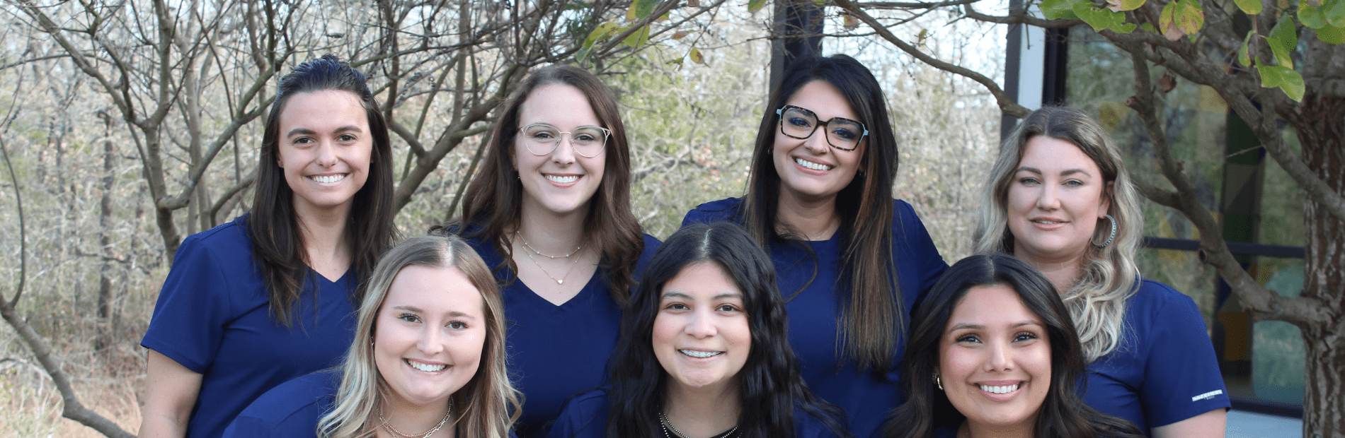 Seven women wearing navy blue tops pose together outdoors in front of trees and a window, all smiling at the camera.