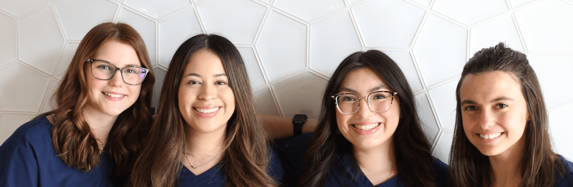 Four young women wearing navy blue tops are smiling in front of a white geometric tiled wall.